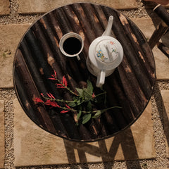 Table basse ronde en bois brûlé Bosc Design vue de dessus, avec cafetière en porcelaine et sauge ananas, sur dalles en pierre
